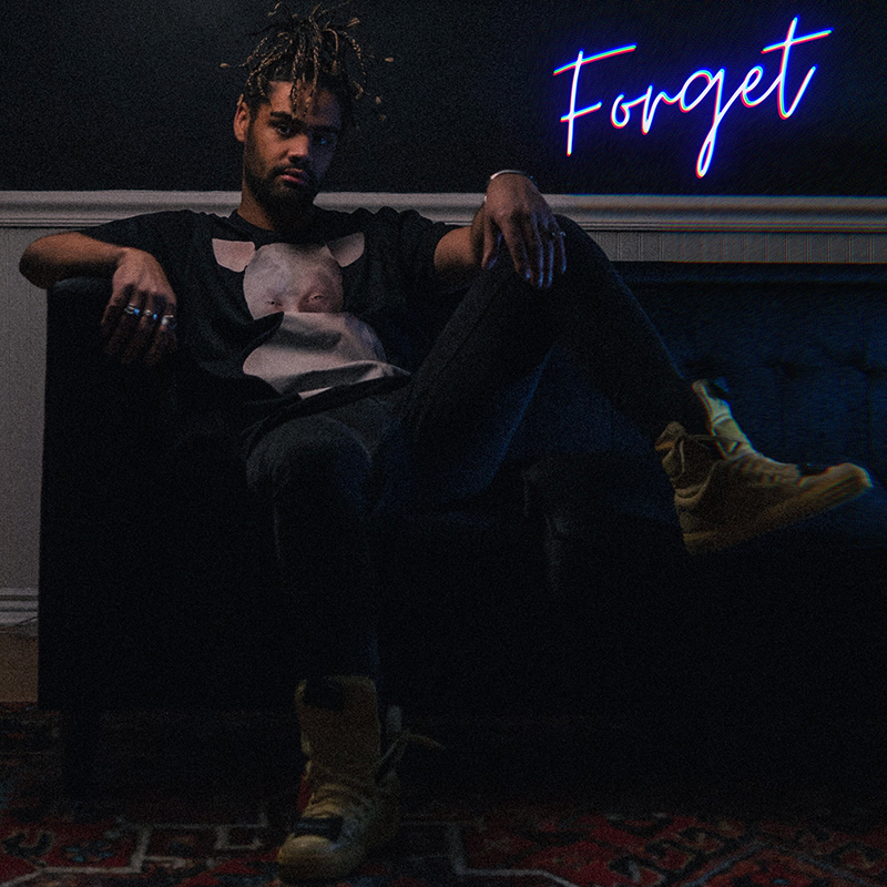 Micah Willis on a couch in dark photograph. In background, a purple neon sign reads "Forget" in cursive.